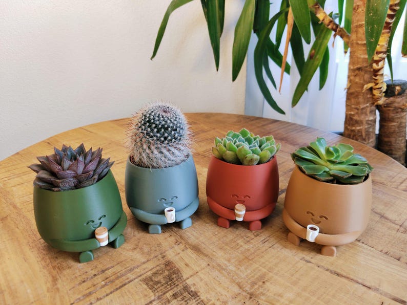 May include: Four colourful, egg-shaped planters with smiling faces, each holding a small coffee cup. The planters are green, blue, red, and tan, and contain succulents and a cactus. The planters are on a wooden table.
