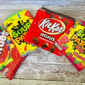 May include: Four colorful zippered pouches with candy bar packaging designs. The pouches are made of fabric and feature the logos for Skittles, Sour Patch Kids, Kit Kat Minis, and Sour Patch Kids.