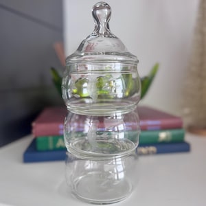 May include: A clear glass apothecary jar with a lid, stacked in three sections. The jar has a decorative design etched into the glass. The jar is sitting on a white surface with books in the background.