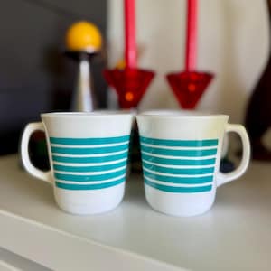 May include: Two white ceramic mugs with teal horizontal stripes. Each mug has a curved handle and a simple, clean design. The mugs are placed on a white surface, with blurred decorative items in the background.