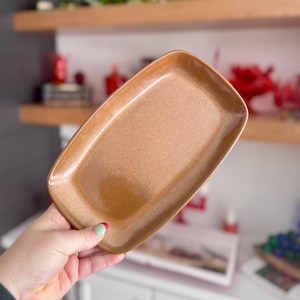 May include: A rectangular serving tray with rounded corners. The tray has a speckled, metallic bronze finish. The tray is being held up in front of a blurred background of shelves and decorative items.