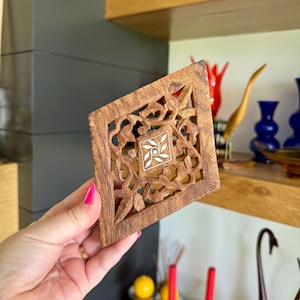 May include: A diamond-shaped, intricately carved wooden coaster with a floral design. The coaster features a central square with a white and cream-colored floral inlay. The wood has a warm, brown tone, and the carving is detailed and delicate.