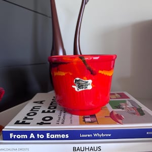 May include: A bright red ceramic planter with a tapered base and a glossy finish, featuring a prominent white sticker label on the front. The planter displays abstract streaks of orange and dark brown glaze across its exterior surface.