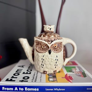 May include: A ceramic owl-shaped teapot with a cream-colored body and brown details. The teapot features a handle, spout, and a small owl figurine on the lid. The teapot is sitting on top of a book titled "From A to Eames".