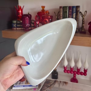 May include: A white, mid-century modern, ceramic serving dish with a unique, asymmetrical shape. The dish is held in a hand and appears to be in excellent condition. The background includes a shelf with books and decorative items.