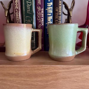 May include: Two ceramic mugs with angular handles and a smooth, glazed finish. The left mug features a gradient of cream and tan, while the right mug displays a soft green hue. Both mugs have a solid, unglazed base and a cylindrical body shape.