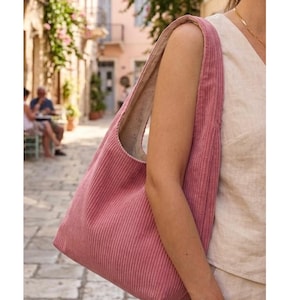 Puede incluir: Un bolso tote de pana rosa polvoriento con una correa ancha para el hombro. El bolso se lleva sobre el hombro. El fondo muestra una escena de calle europea con edificios y personas.