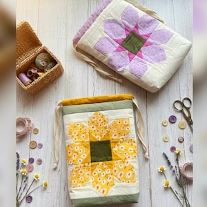 May include: Two handmade fabric bags with floral designs. One bag is a drawstring style with a yellow flower pattern, and the other is a rectangular bag with a purple flower design. A woven basket with spools of thread and sewing notions is also visible.