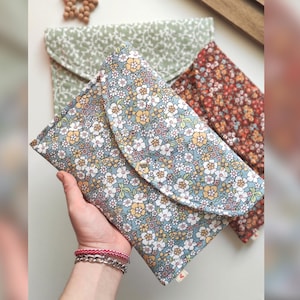 May include: Three floral print pouches in blue, green, and brown. The front pouch is held by a hand and features a blue background with small white, yellow, and orange flowers. The other two pouches have similar floral patterns.