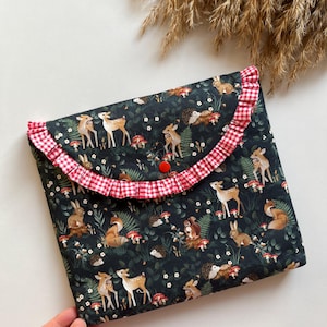 May include: A dark green fabric case with a woodland animal print featuring deer, foxes, rabbits, and mushrooms. The case has a red and white gingham ruffle trim and a red button closure.