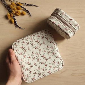 Puede incluir: Dos neceseres con estampado floral crema con flores rosas y rojas. Uno es rectangular con cremallera, el otro es cuadrado y abierto. Flores amarillas y moradas secas en el fondo.