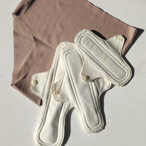 Könnte beinhalten: Drei weiße wiederverwendbare Stoff-Menstruationseinlagen auf einem braunen Stoffunterwäsche.