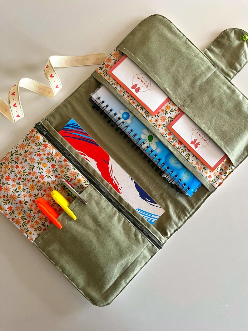 Puede incluir: Un organizador de tela verde oliva abierto con forro estampado floral. Contiene cuadernos, tarjetas y bol&iacute;grafos. Se ve una cinta blanca con corazones rojos. El organizador tiene cierre de cremallera y bot&oacute;n.