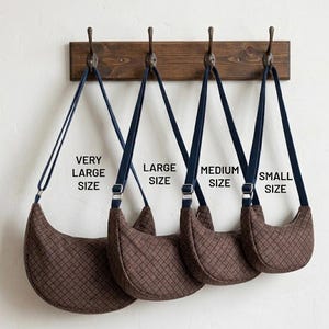 Könnte beinhalten: Vier braune, halbmondförmige Taschen in verschiedenen Größen, beschriftet mit "Very Large Size", "Large Size", "Medium Size" und "Small Size". Jede Tasche hat einen dunkelblauen Riemen und hängt an einer Holzleiste mit Metallhaken.
