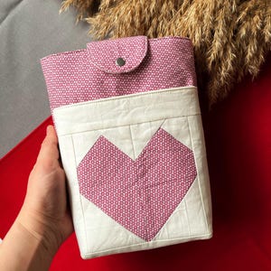 May include: A handmade, quilted book cover with a heart design. The cover features a pink and white geometric pattern, a large heart applique, and a snap closure. The background is red and gray.