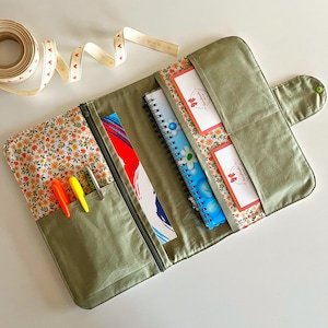 Puede incluir: Organizador de tela verde oliva abierto con forro estampado floral. El organizador tiene un bolsillo con cremallera con bol&iacute;grafos, un bolsillo con un cuaderno y un bolsillo con dos cuadernos. Un rollo de cinta con detalles de coraz&oacute;n est&aacute; en el fondo.