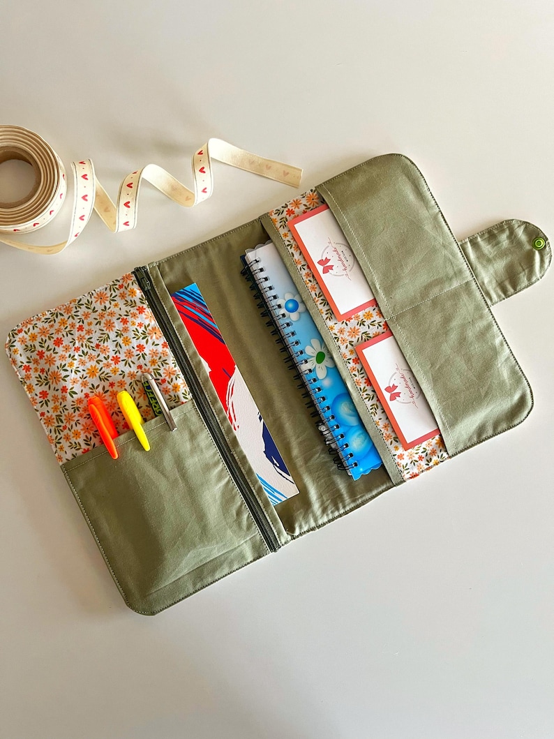 Puede incluir: Organizador de tela verde oliva abierto con forro estampado floral. El organizador tiene un bolsillo con cremallera con bol&iacute;grafos, un bolsillo con un cuaderno y un bolsillo con dos cuadernos. Un rollo de cinta con detalles de coraz&oacute;n est&aacute; en el fondo.