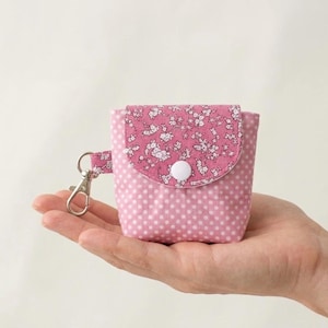 Könnte beinhalten: Kleine, rosa Stofftasche mit weißen Punkten und floralem Klappverschluss. Die Tasche hat einen silberfarbenen Verschluss und einen weißen Knopfverschluss. Die Tasche wird in einer Hand gehalten.