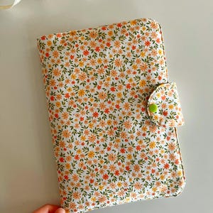 Puede incluir: Una funda de libro con estampado floral con fondo blanco y flores naranjas y amarillas. La funda tiene un cierre de bot&oacute;n verde. Una cinta blanca con corazones rojos est&aacute; en el fondo.