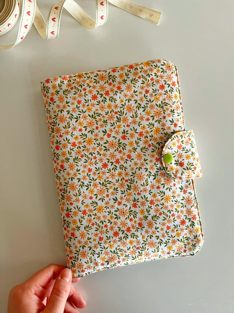 Puede incluir: Una funda de libro con estampado floral con fondo blanco y flores naranjas y amarillas. La funda tiene un cierre de bot&oacute;n verde. Una cinta blanca con corazones rojos est&aacute; en el fondo.