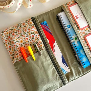 Puede incluir: Un organizador de tela verde oliva abierto con cremallera, que revela un interior con estampado floral y bolsillos. En el interior hay bol&iacute;grafos, cuadernos y un marcap&aacute;ginas. Se ve un rollo de cinta con detalles de coraz&oacute;n.