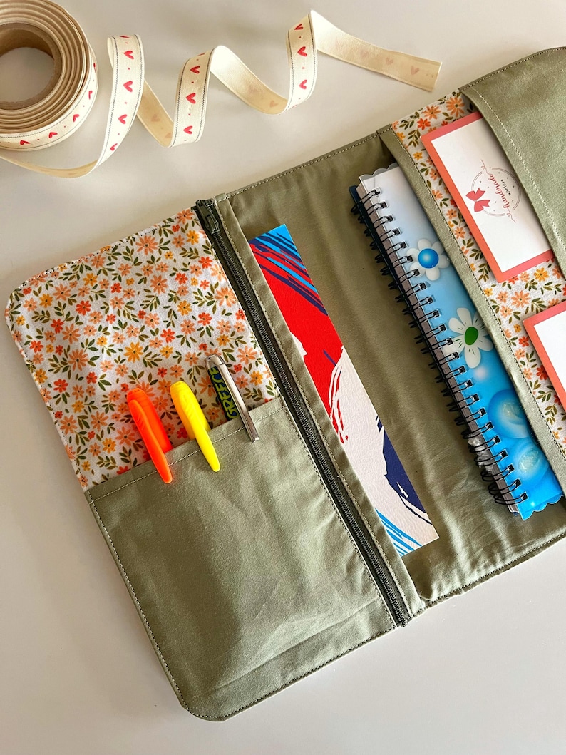 Puede incluir: Un organizador de tela verde oliva abierto con cremallera, que revela un interior con estampado floral y bolsillos. En el interior hay bol&iacute;grafos, cuadernos y un marcap&aacute;ginas. Se ve un rollo de cinta con detalles de coraz&oacute;n.