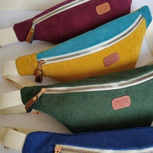 Peut inclure: Plusieurs sacs banane en velours côtelé de couleurs bordeaux, turquoise, jaune, vert et bleu. Chaque sac est doté d'une fermeture éclair et d'une sangle contrastantes, avec une petite étiquette en cuir. Les sacs sont de forme incurvée.