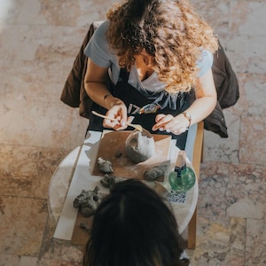 May include: An overhead shot of a person sculpting clay on a small round table. The person is wearing a black apron and using tools to shape the clay. Another person is seated in front, observing the process. A green bottle and clay pieces are also on the table.