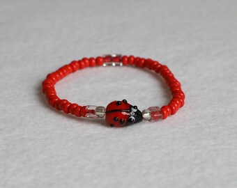Könnte beinhalten: Rotes Perlenarmband mit einem Marienkäfer-Anhänger. Der Marienkäfer ist aus Glas und hat schwarze Punkte.