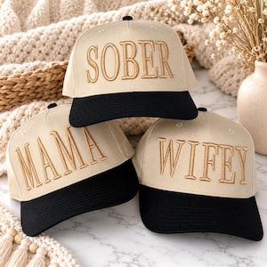May include: Three baseball caps with a cream-colored crown and a black brim. Each cap features embroidered gold lettering on the front. The top cap reads SOBER, the bottom left cap reads MAMA, and the bottom right cap reads WIFEY in a serif font.