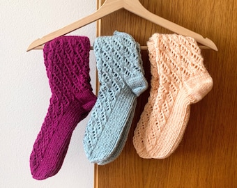 May include: Three pairs of knitted socks in shades of purple, light blue, and peach, hanging on a wooden hanger. Each sock features a diamond-shaped openwork pattern. The socks are displayed against a wooden door.