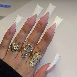 May include: A hand displaying long, square-shaped acrylic nails with a white French tip design. The nails have a pink base. The hand also features several rings, including a gold heart-shaped ring and a gold rectangular ring with a figure.