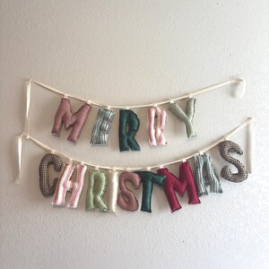 May include: A festive "MERRY CHRISTMAS" banner. The letters are made of fabric in various patterns and colors, including red, green, and plaid. The banner is strung on a cream-colored ribbon and is hanging against a white wall.