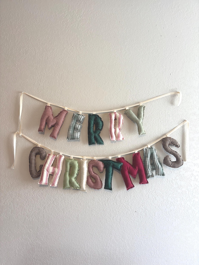 May include: A festive "MERRY CHRISTMAS" banner. The letters are made of fabric in various patterns and colors, including red, green, and plaid. The banner is strung on a cream-colored ribbon and is hanging against a white wall.