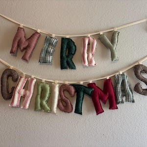 May include: A fabric banner spelling "MERRY CHRISTMAS" in various patterned letters. The letters are in red, green, and white fabrics, suspended from a cream-colored ribbon. A festive, handmade decoration for the holiday season.