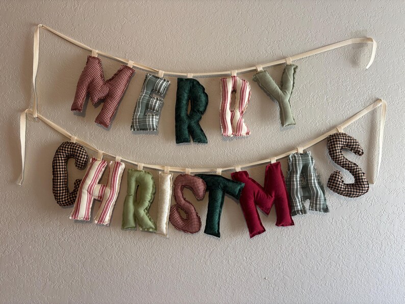 May include: A fabric banner spelling "MERRY CHRISTMAS" in various patterned letters. The letters are in red, green, and white fabrics, suspended from a cream-colored ribbon. A festive, handmade decoration for the holiday season.