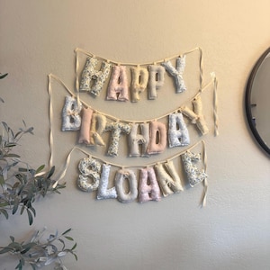 May include: A fabric banner featuring three rows of stuffed, plush letters spelling out HAPPY BIRTHDAY SLOANE. The letters are crafted from various pastel-colored fabrics with floral and striped patterns, suspended on thin, light-colored ribbon strings.