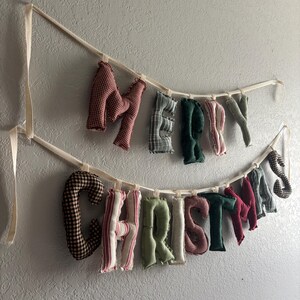 May include: A fabric banner spelling "MERRY CHRISTMAS" in various patterns and colors. The letters are stuffed and sewn, with a mix of red gingham, green, and striped fabrics. The banner hangs from a cream-colored ribbon against a textured wall.