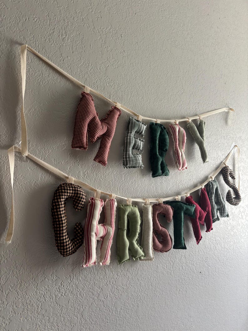 May include: A fabric banner spelling "MERRY CHRISTMAS" in various patterns and colors. The letters are stuffed and sewn, with a mix of red gingham, green, and striped fabrics. The banner hangs from a cream-colored ribbon against a textured wall.