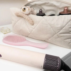 May include: A beige and white gingham quilted toiletry bag with a plush bear toy attached to the front. Inside the bag are hair styling attachments. A light pink hairbrush and a long, cylindrical hair styling tool with a dark gray mesh end.