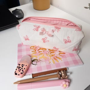 Può includere: Un astuccio per matite trapuntato rosa e bianco con una fodera a quadri rosa. L'astuccio è decorato con fiocchi rosa e ha una chiusura a zip. L'astuccio è appoggiato su un quaderno a quadri rosa e bianco con le parole "sii gentile" scritte in glitter dorato. Ci sono anche diverse penne e matite sul quaderno.
