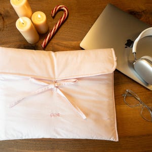 May include: A pink fabric pouch with a tied ribbon and the word "GOA" embroidered on the front. The pouch is on a wooden surface with a laptop, headphones, glasses, and a candle.