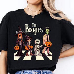 May include: Black t-shirt featuring a Halloween-themed graphic of four characters crossing a street, each holding a jack-o'-lantern. The characters are dressed as a witch, a boy, a mummy, and Frankenstein's monster. The text "The Bootles" is above them.