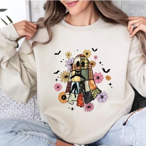 May include: Cream-colored sweatshirt featuring a patchwork ghost design with a black cat, surrounded by flowers and bats. The ghost has a stitched appearance, with a variety of colorful fabric squares. A fun, festive design for Halloween.