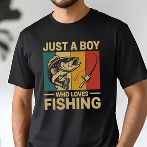 May include: Black t-shirt with a graphic featuring a fish and fishing rod. The text reads "JUST A BOY WHO LOVES FISHING" in a vintage style. The design incorporates teal, yellow, and red colors.
