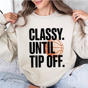 May include: Cream-colored sweatshirt with the text "CLASSY. UNTIL TIP OFF." in black, and an orange basketball graphic. The sweatshirt is a casual style, suitable for sports fans.