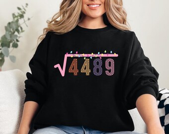 May include: A black long-sleeve sweatshirt featuring a graphic of a square root symbol over the number 4489. The numbers have a checkered pattern, and the square root bar is decorated with colorful, glowing Christmas light bulbs.