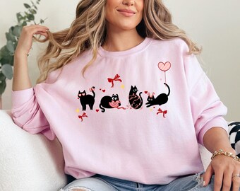 May include: A light pink sweatshirt featuring a playful design of four black cats. Each cat is engaged in a different activity, such as holding a heart-shaped balloon or playing with yarn. Red bows and hearts add to the festive theme.