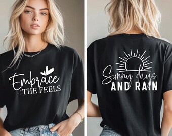 May include: Black t-shirt with white text that says "Embrace the Feels" on the front and "Sunny days AND RAIN" on the back. The front design includes a heart and leaf graphic. The back design features a sun graphic.