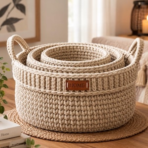 May include: A set of three nested round baskets made of thick beige rope with a textured crochet-style weave. Each basket features two integrated side handles and a small rectangular brown leather-like patch with the word HOME embossed on it.